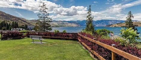 A grassy area with a wooden fence, a bench, and a view of a lake with mountains in the background.