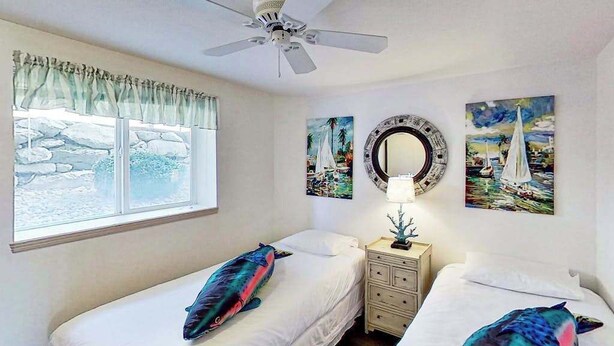 Quarto com duas camas, um ventilador de teto, janela com vista para rochas e obras de arte nas paredes.