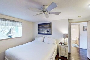 Ein Schlafzimmer mit Bett, Deckenventilator, Gemälde, Nachttisch und einer Tür zu einem Badezimmer.