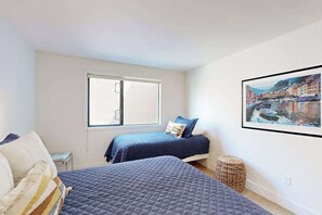 Ein Schlafzimmer mit zwei Betten, einem Fenster, einem gerahmten Bild an der Wand und einem kleinen Korbsessel.