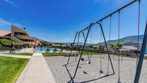 A playground with swings and a pool area, surrounded by a building with a sloped roof. - 2BR Lakefront - Patio - Pool - Tennis - Dock (Chelan)