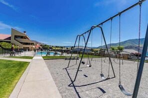 A playground with swings and a pool area, surrounded by a building with a sloped roof. - 2BR Lakefront - Patio - Pool - Tennis - Dock (Chelan)