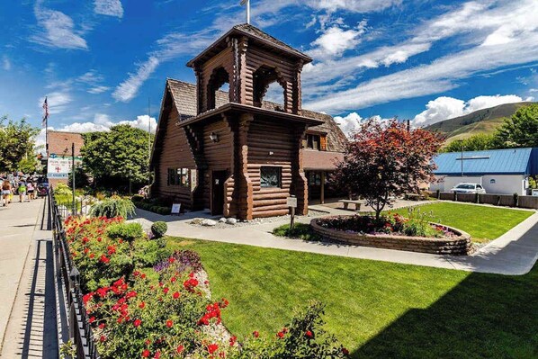Uma cabana de madeira com um campanário, rodeada por um jardim bem cuidado com diversas plantas e flores.