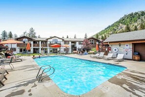 A swimming pool with lounge chairs and umbrellas, surrounded by buildings with a mountain backdrop. - 2BR 1st-Floor - Icicle Village Resort Condo (Leavenworth)
