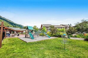 Un parque infantil con toboganes y aro de baloncesto en un área verde, con edificios al fondo.
