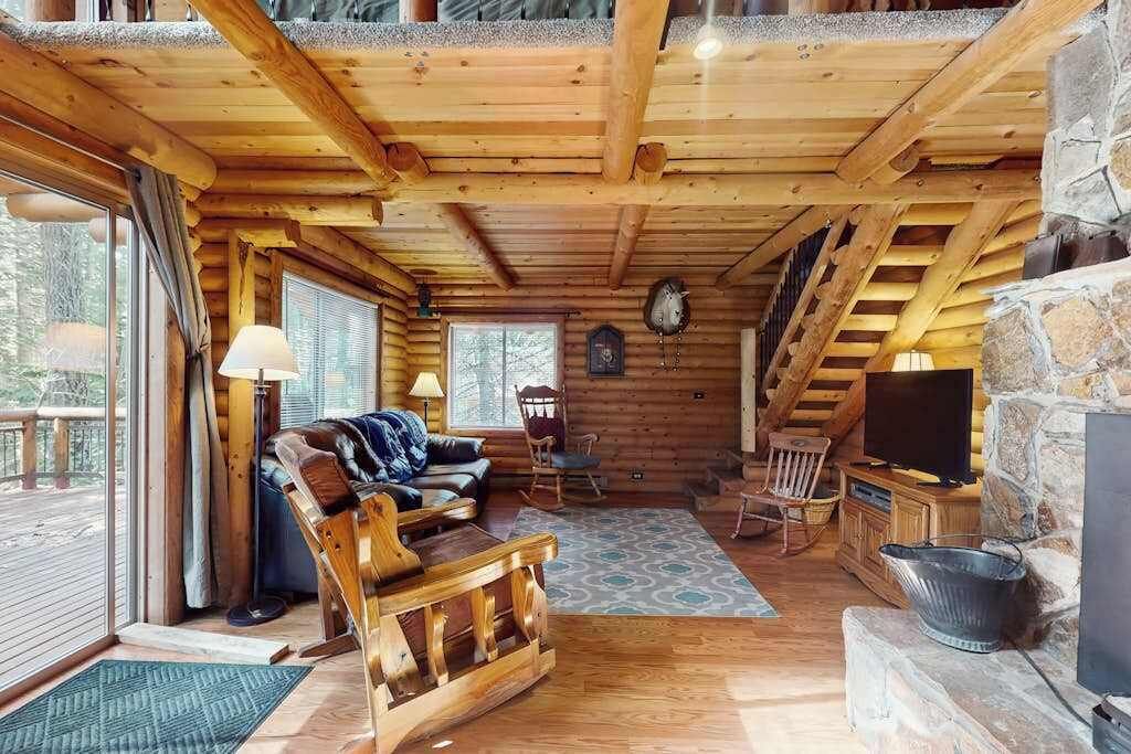 A log cabin interior with a wooden ceiling, a stone fireplace, a television, and a cozy seating area with a sofa and chairs.