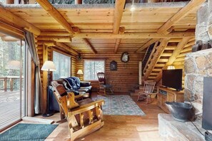 Interior de una cabaña de troncos con techo de madera, chimenea de piedra, televisor y una zona de estar acogedora con sofá y sillones.