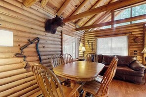 Ein rustikales Blockhaus-Interieur mit Esstisch, Stühlen, einer Couch und einem aufgehängten Bearranhänger.