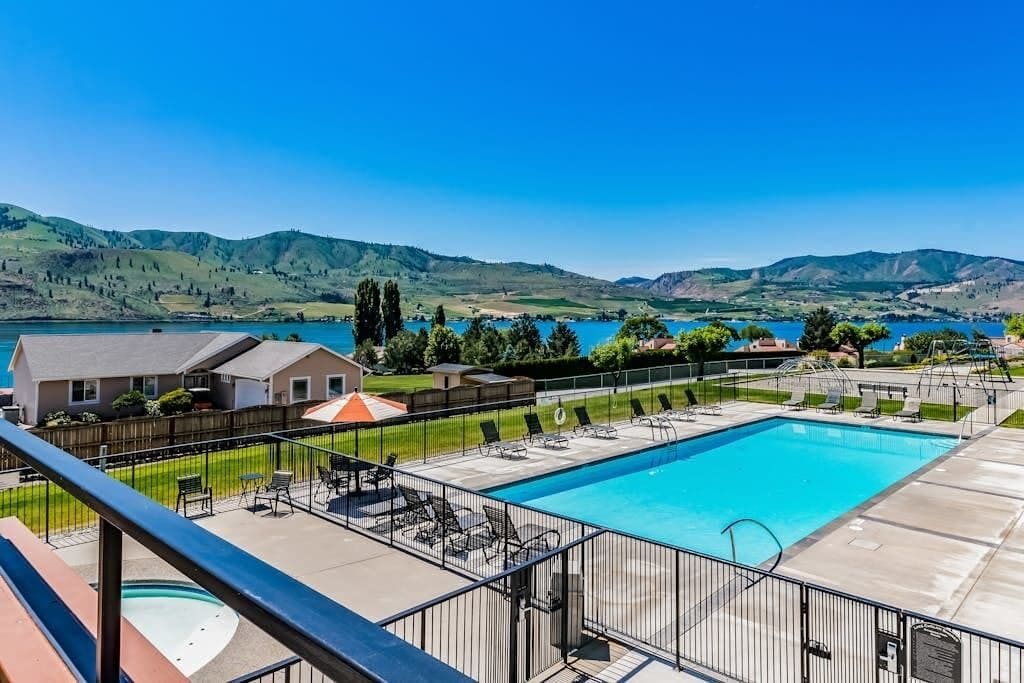 Ein Swimmingpool mit Blick auf Berge und einen See, umgeben von einer eingezäunten Terrassenanlage mit Liegestühlen.