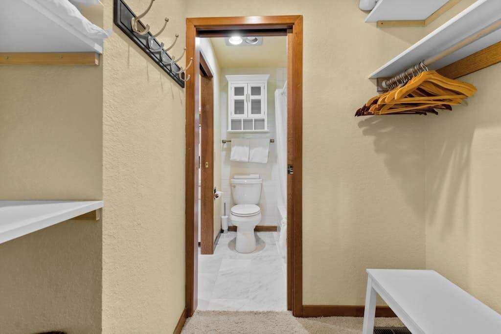 Een smalle gang die uitkomt bij een badkamer, met een toilet, een witte handdoekhouder en een witte plank met kledinghangers.