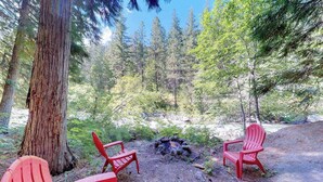 Zona boscosa con una hoguera y tres sillas Adirondack rojas.