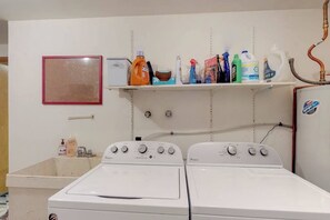 A laundry area with two front-loading washing machines, a shelf with cleaning supplies, and a red-framed picture on the wall.