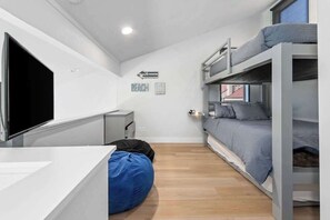 Quarto compacto com beliches, uma escrivaninha, um puf e uma televisão.