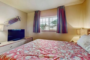 A bedroom with a bed, a television on a stand, a window with curtains, and a view of houses outside. - 2 BR condos located in the Icicle Resort (Leavenworth)