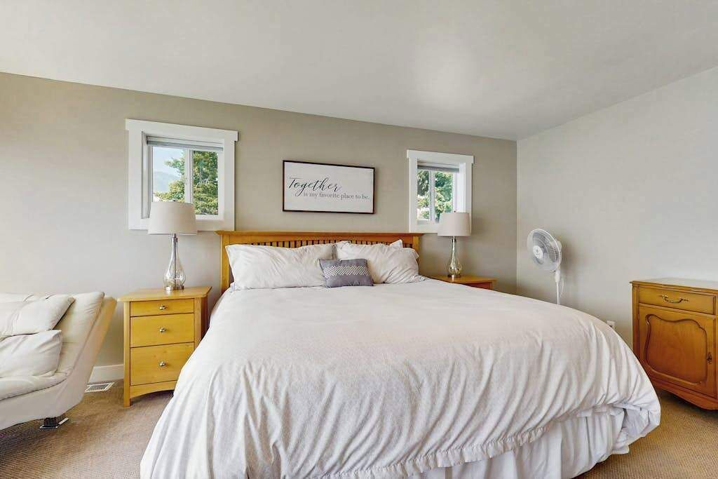 A bedroom with a large bed, two bedside tables with lamps, a wall-mounted fan, and a framed quote above the bed.