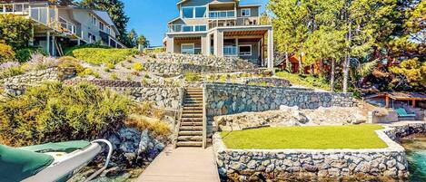 A coastal property with a wooden deck leading to a stone staircase, a clear blue pool, and a two-story house with a balcony.