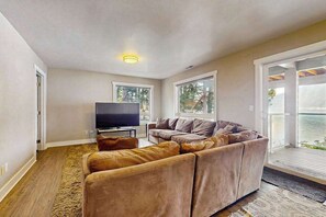 Ein Wohnzimmer mit einem Ecksofa, einem Flachbildfernseher und großen Fenstern mit Blick ins Freie.