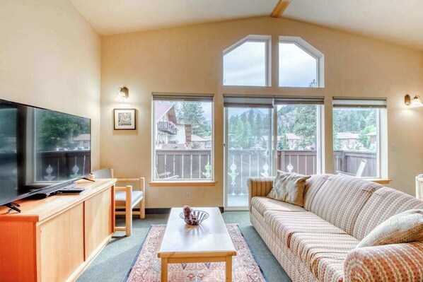 A living room with a television, sofa, and a view of a deck and trees. - Downtown 2BR Mountainview 2nd-Floor - Balcony (Leavenworth)