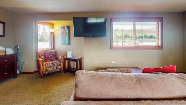 A bedroom with a bed, a TV mounted on the wall, a window with blinds, a chair, and a small table. - Lovely Studio Mountainview - Patio - Pool (Chelan)