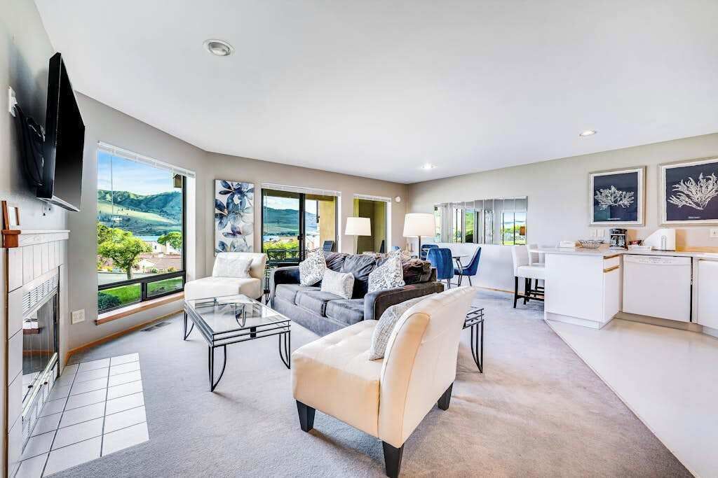 A spacious living room with a sofa, armchairs, and a coffee table. There is a kitchen area with a white countertop and cabinets.