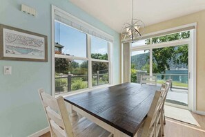 A dining area with a long table, white chairs, and large windows offering a view of greenery and a body of water. - Hip 4BR Lakeview - Pool - Hot Tub - Deck (Manson)