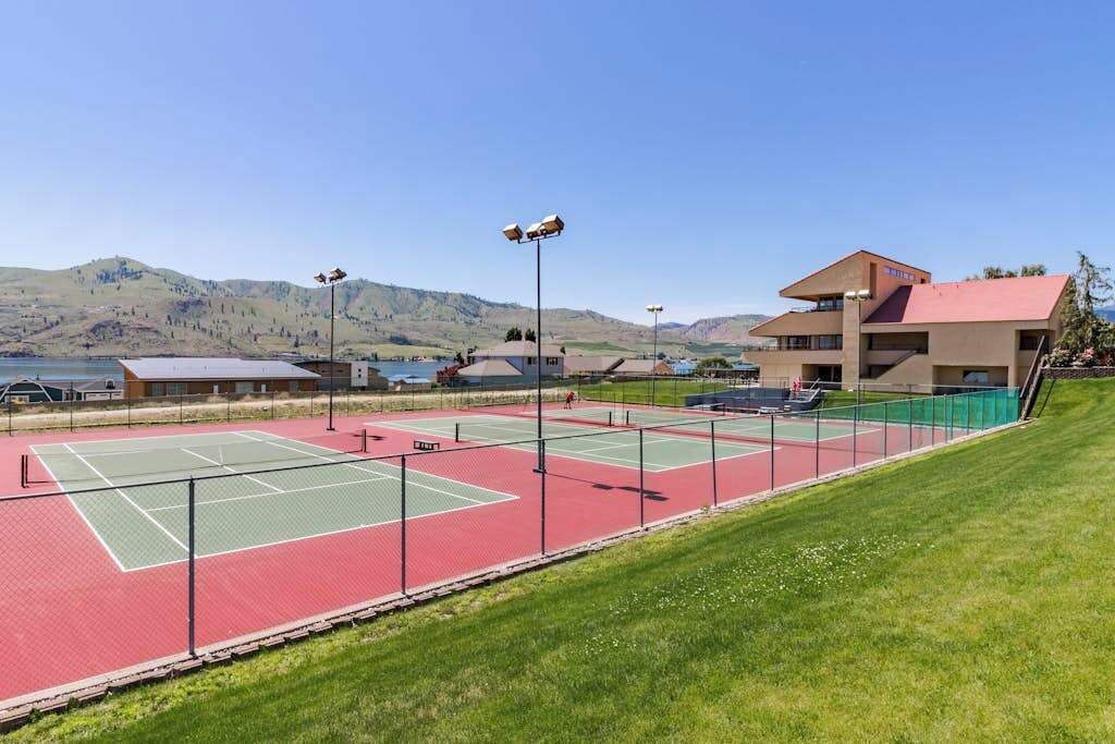 A tennis court with a red and green surface, surrounded by a fence, with a building and mountains in the background.