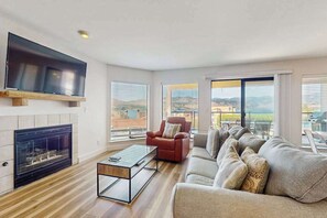 A modern living room with a fireplace, a flat-screen TV mounted above it, a red armchair, a beige sofa, and large windows offering a view of the outdoors. - Large 2BR Lakeview 1st-Floor - Balcony - Pool (Chelan)