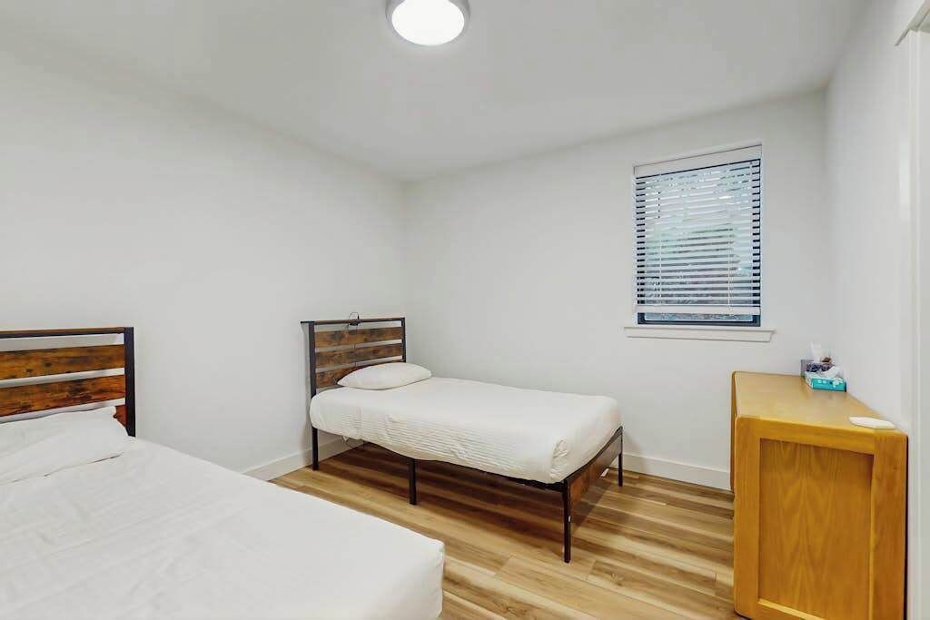 A bedroom with two beds, a wooden floor, a window with blinds, and a small wooden table.