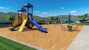 Ein Spielplatz mit Rutsche und Klettergerüsten, umgeben von einer angelegten Grünfläche mit Bänken und Blick auf Hügel.