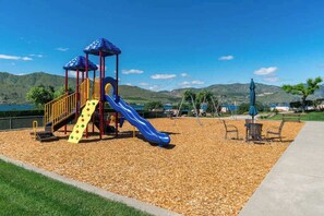 A playground with a slide and climbing structures, surrounded by a landscaped area with benches and a view of hills. - Large 2BR Lakeview 1st-Floor - Balcony - Pool (Chelan)