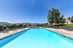 A rectangular swimming pool with a clear blue water surface, surrounded by lounge chairs and umbrellas, with a view of hills in the background. - 2BR Lakeview 1st-Floor - Deck - Pool (Chelan)