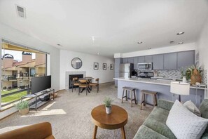 Ein modernes Wohnzimmer mit einer Couch, einem Couchtisch und einem Fernseher. Es gibt eine Küchenzeile mit einer Mikrowelle und einem Esstisch.