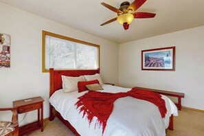 Ein Schlafzimmer mit Bett, Nachttisch, Sitzbank, Deckenventilator, Fenster mit Jalousien und einem gerahmten Bild an der Wand.