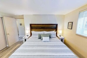 Une chambre d’hôtel avec un grand lit, deux tables de chevet, une tête de lit en bois et une fenêtre avec des stores.