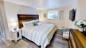 A bedroom with a wooden headboard, a bed with striped bedding, a bedside table with a lamp, a small chair, a dresser, and a TV mounted on the wall. - Spacious 2BR Mountainview 1st-floor - Patio (Chelan)