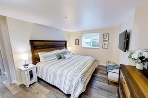Ein Schlafzimmer mit einem hölzernen Kopfteil, einem Bett mit gestreifter Bettwäsche, einem Nachttisch mit Lampe, einem kleinen Stuhl, einer Kommode und einem an der Wand montierten Fernseher.