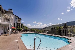 A swimming pool with a metal ladder, surrounded by a concrete deck, with a mountain backdrop and a building with a red awning.