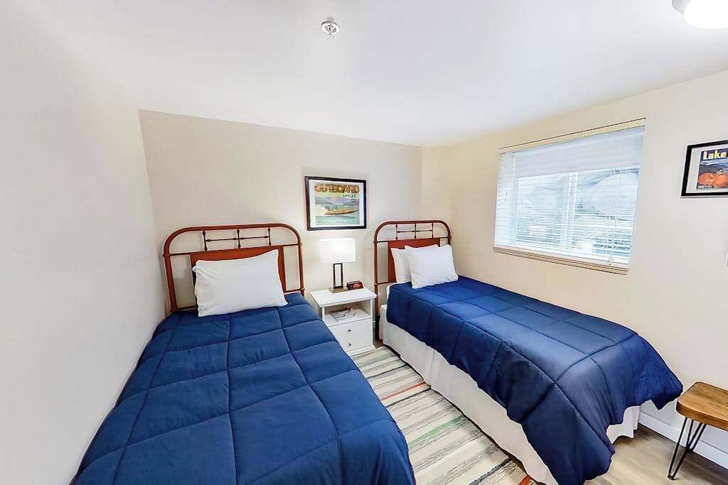 A room with two single beds, a small bedside table with a lamp, a framed picture on the wall, and a window with blinds.