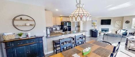 A modern kitchen with a central island, dining area with a table and chairs, and a living area with a sofa and TV.