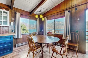 A dining area with a round table and wooden chairs, large windows with curtains, and a view of a mountainous landscape.