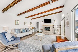 Un salon spacieux avec un canapé blanc, un fauteuil bleu et une table basse ronde. On aperçoit une cheminée au fond de la pièce, et des tableaux encadrés ornent les murs.