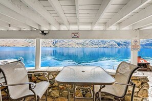 A covered outdoor seating area with a table and chairs, overlooking a body of water with a mountainous backdrop.