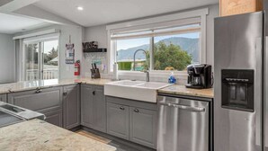 A modern kitchen with grey cabinets, a stainless steel refrigerator, a built-in dishwasher, and a window with a view of greenery and mountains. - Walk to Downtown 3BR Lakeview - Hot Tub - Deck (Manson)