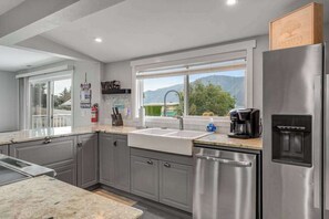 A modern kitchen with grey cabinets, a stainless steel refrigerator, a built-in dishwasher, and a window with a view of greenery and mountains. - Walk to Downtown 3BR Lakeview - Hot Tub - Deck (Manson)