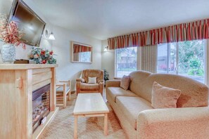 Ein Wohnzimmer mit Sofa, Sessel, Couchtisch, Kamin, Fernseher und Fenstern mit Vorhängen.