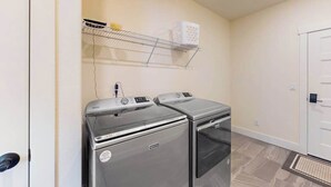 A laundry room with two front-loading washing machines, a metal shelf above, and a door to the right. - 3BR water view home with deck & open layout (Manson)