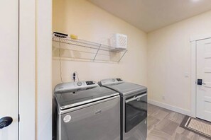 A laundry room with two front-loading washing machines, a metal shelf above, and a door to the right. - 3BR water view home with deck & open layout (Manson)