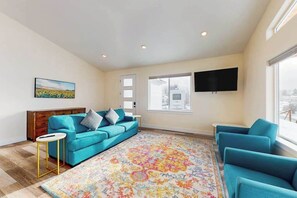 Un salon avec un canapé bleu, un tapis à motifs, un téléviseur à écran plat fixé au mur et une fenêtre donnant sur l’extérieur.