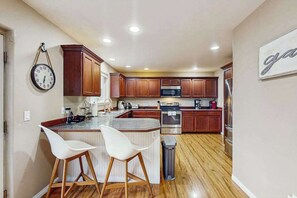 Eine Küche mit Holzausstattung, einem Edelstahlkühlschrank, einer Mikrowelle und einem Spülbecken. Zwei weiße Barhocker und eine Uhr sind an der Wand angebracht.
