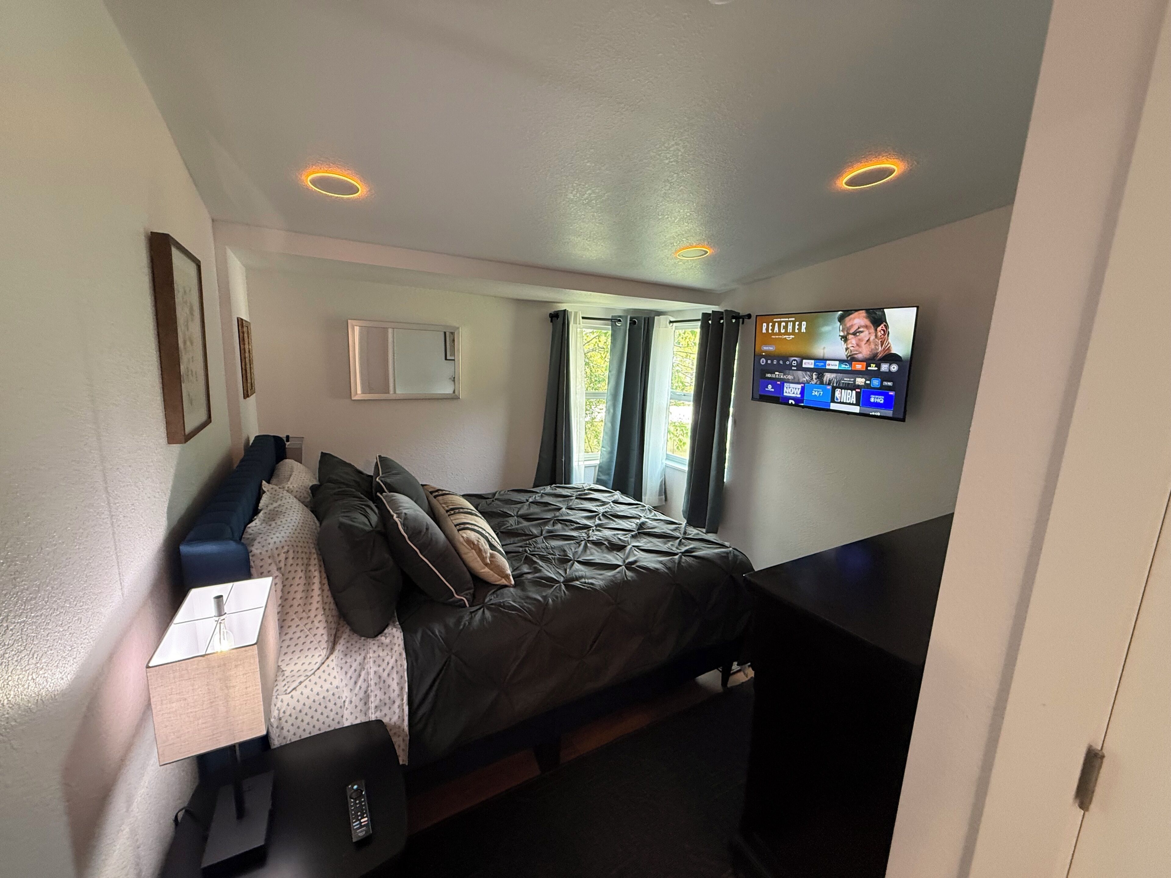 Ein Schlafzimmer mit einem Bett, einem an der Wand montierten Fernseher, einem Nachttisch und einem Fenster mit Vorhängen.